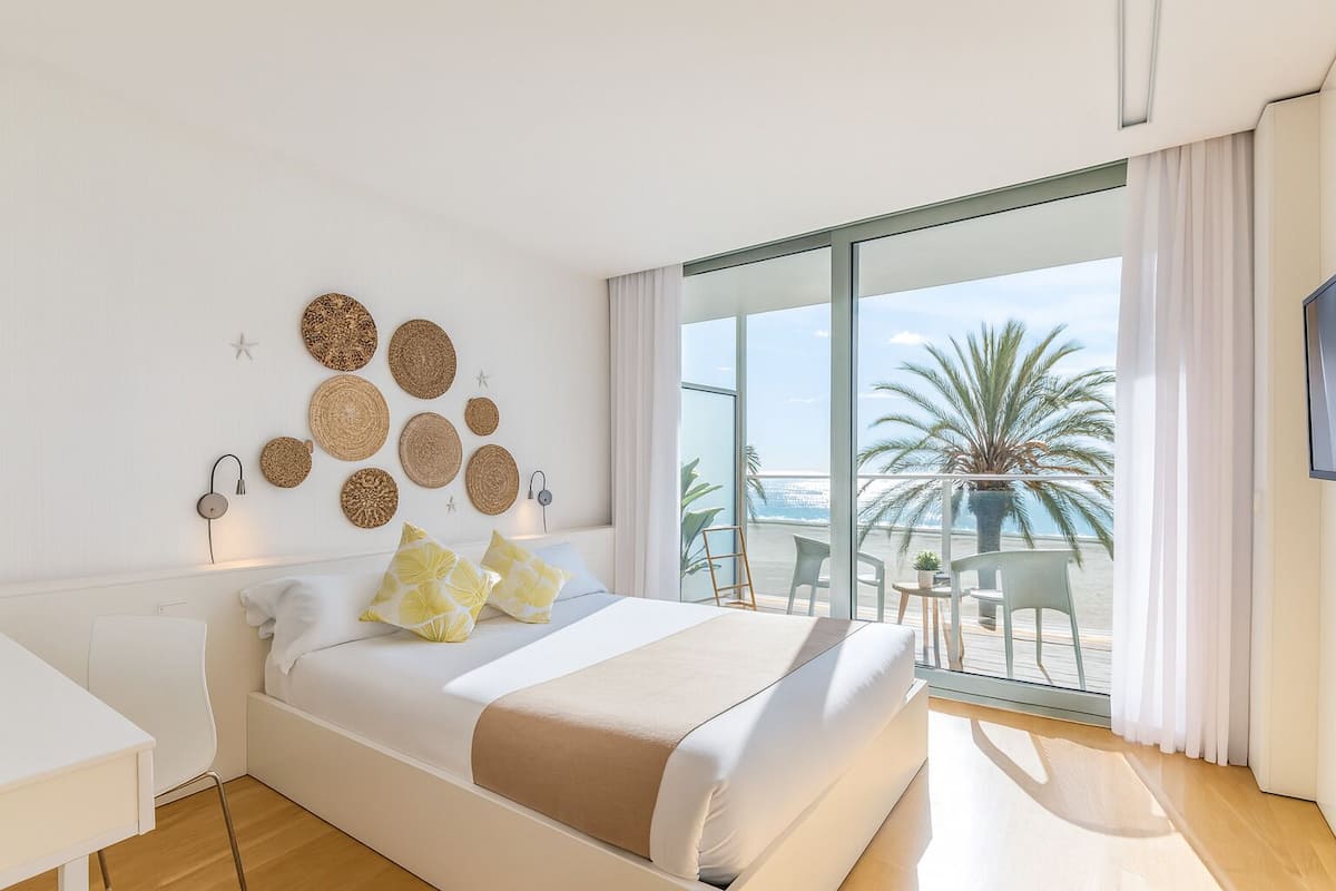 A modern hotel room with a large bed, a balcony with a view of the sea, and decorative woven wall art.