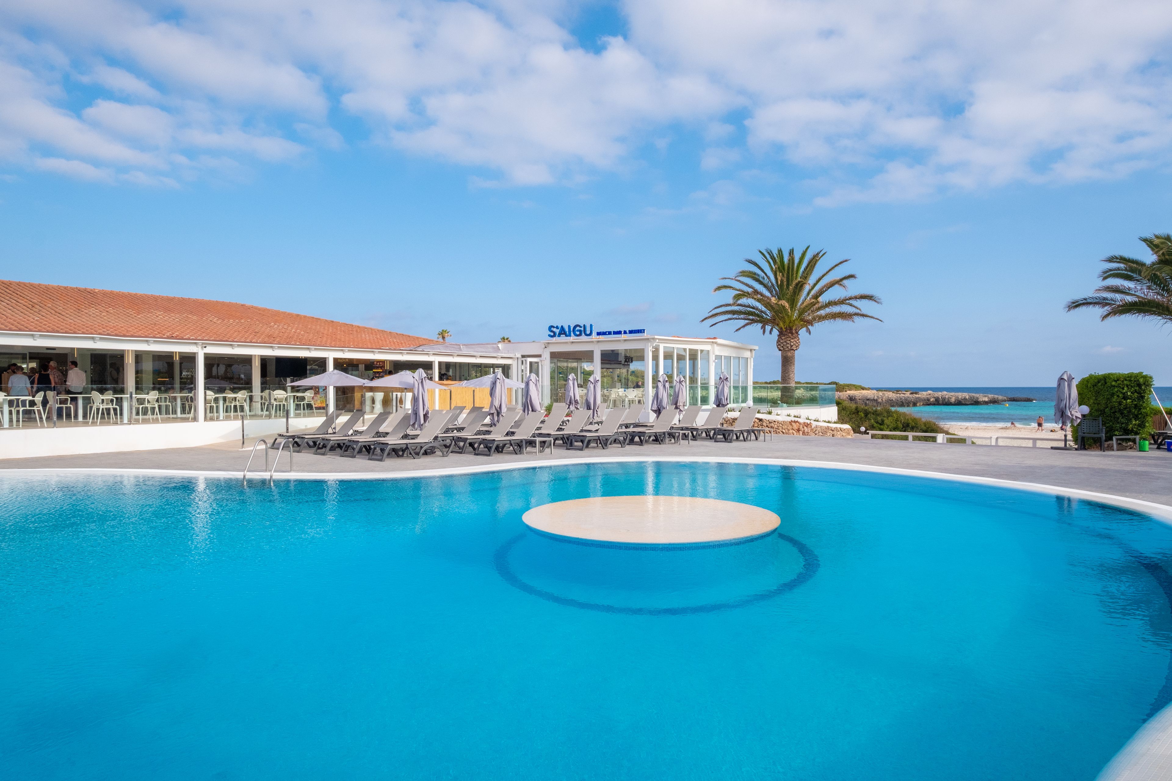 A swimming pool with lounge chairs and umbrellas, a building with a sign reading "SAIGU", and a palm tree.