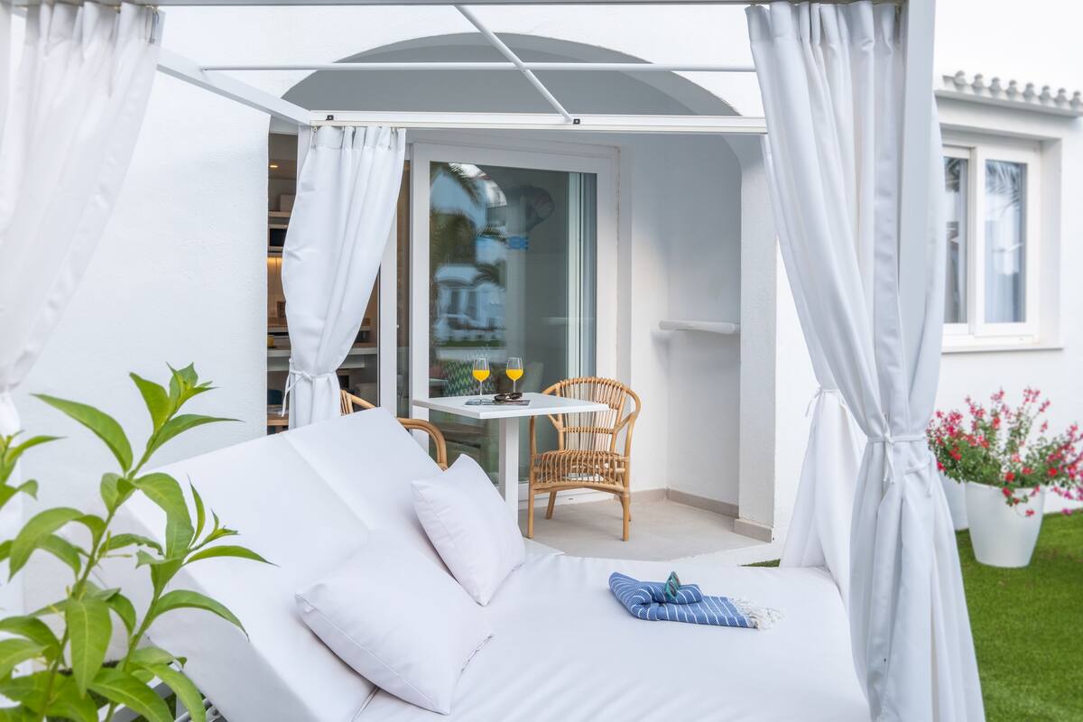 Un área de estar exterior blanca con una mesa y sillas, un porche cubierto y una casa al fondo.