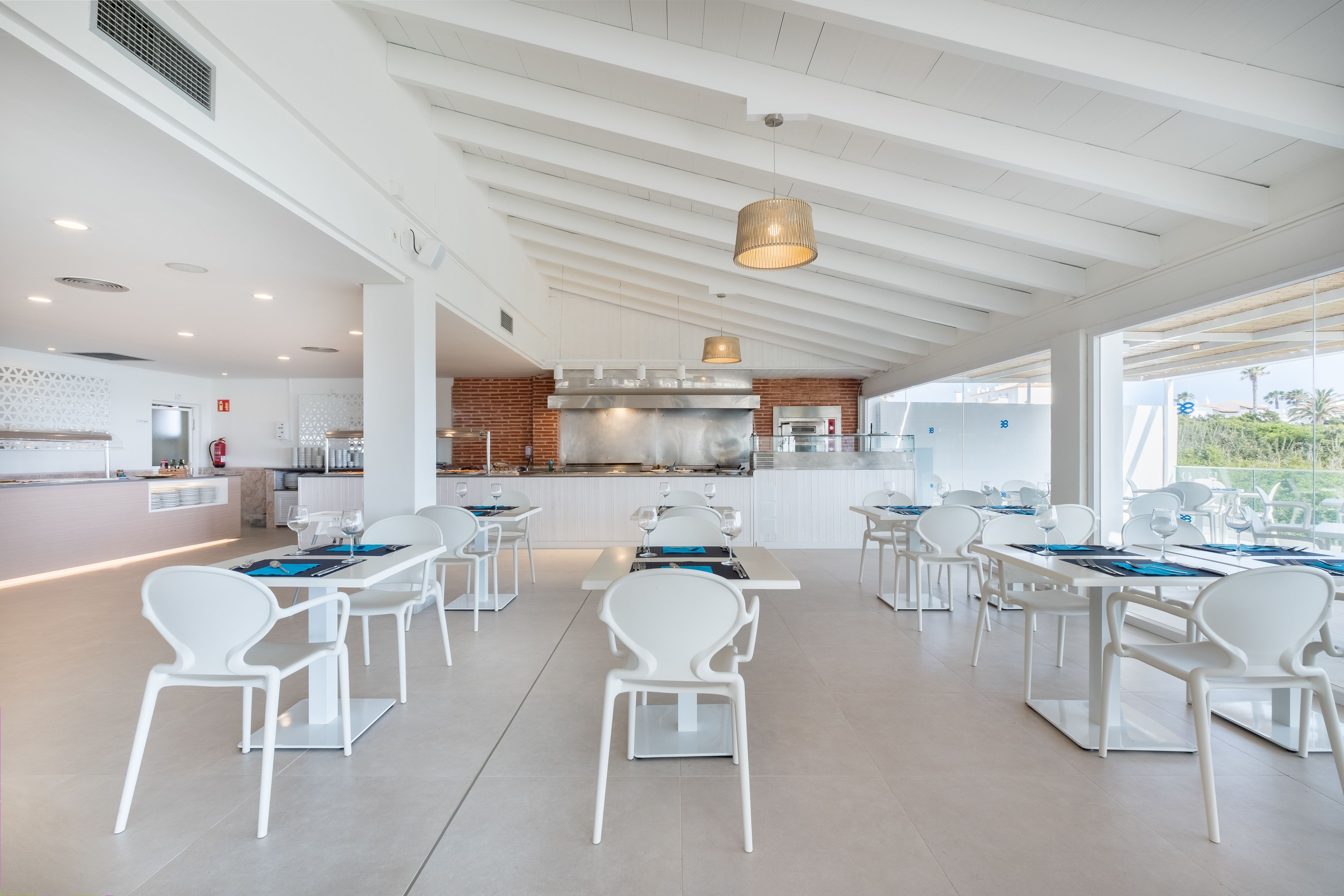 Un restaurante moderno con sillas y mesas blancas, una cocina central y amplios ventanales que dan a vistas del exterior.