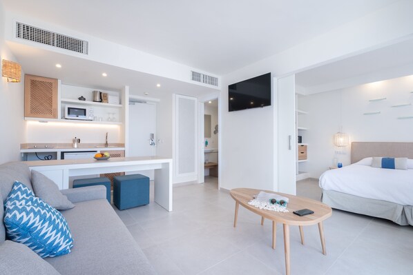 A modern hotel room with a sofa, a dining table, a bed, a TV, and a kitchenette.