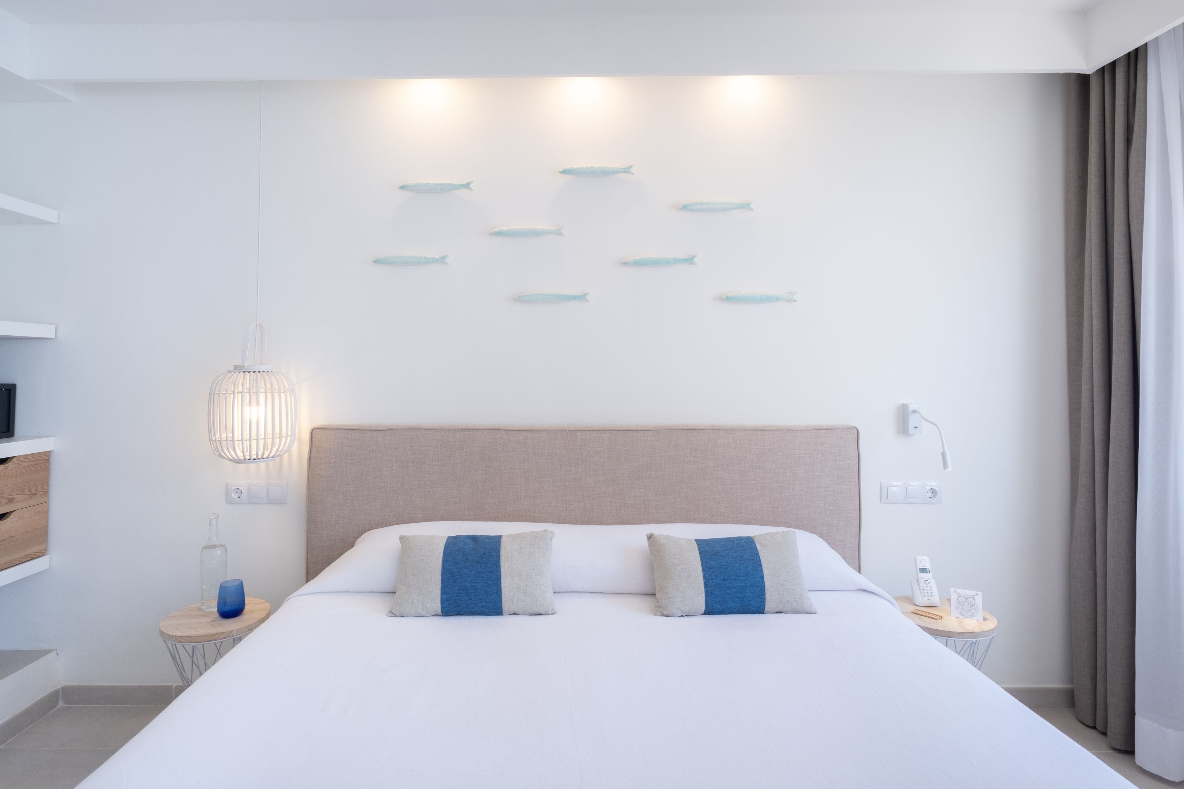 A modern hotel room with a large bed, bedside tables, a wall-mounted light fixture, and a view of the sea.