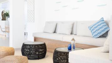 Un salón moderno con un sofá blanco, dos otomanas de mimbre negro y una mesa de centro de vidrio con cristalería azul y transparente.