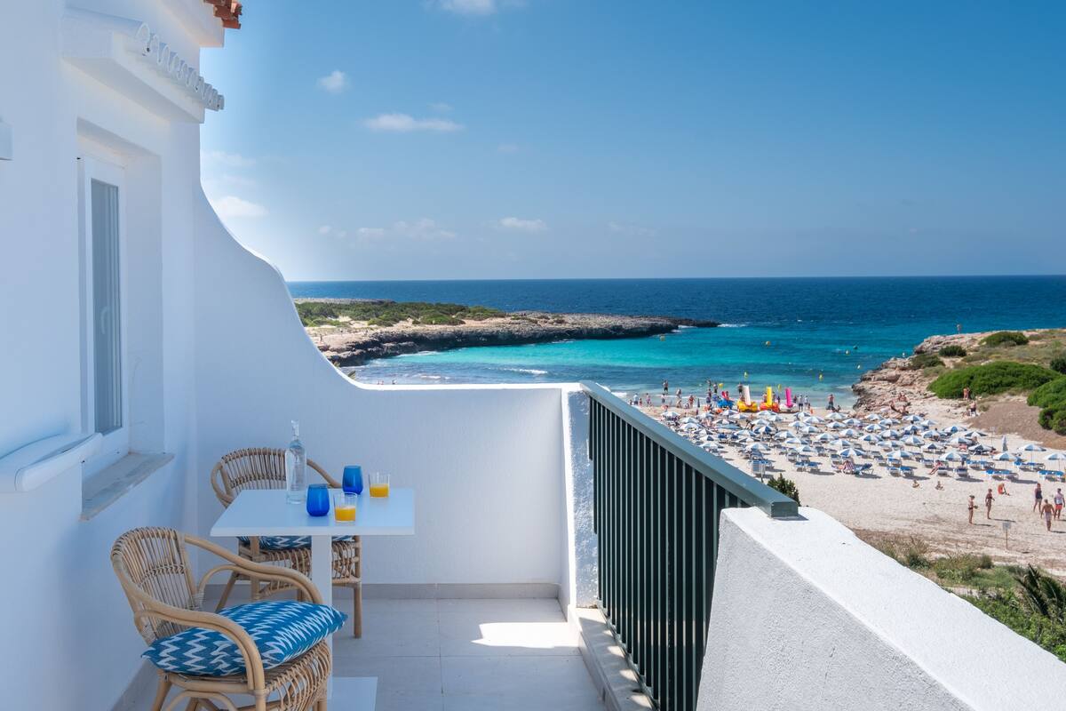 Un balcón con sillas de mimbre y una mesa, con vistas a una playa con sombrillas y gente.