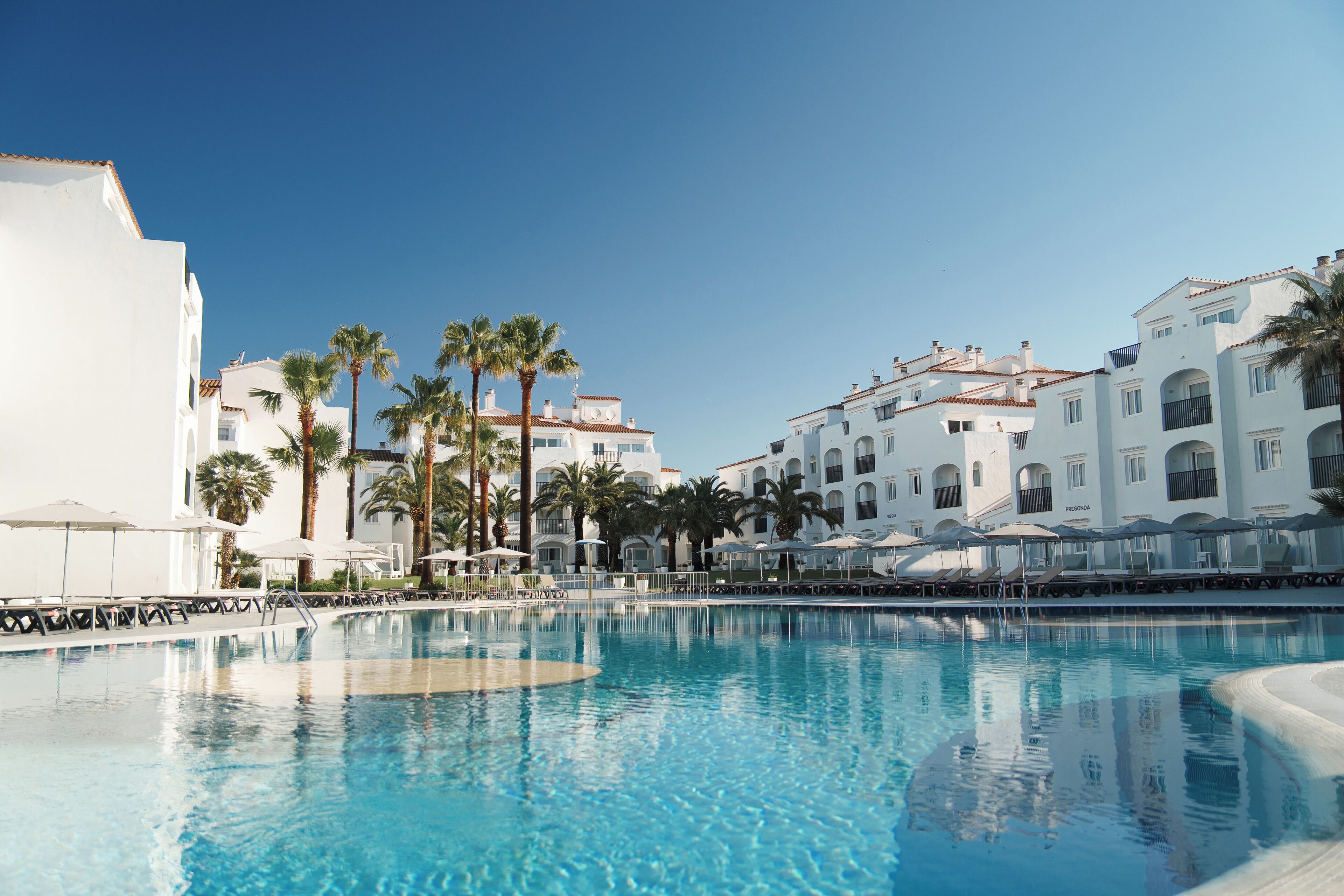 A swimming pool with lounge chairs and umbrellas, surrounded by white buildings with balconies and palm trees.