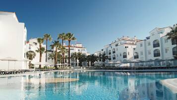 A swimming pool with lounge chairs and umbrellas, surrounded by white buildings with balconies and palm trees.