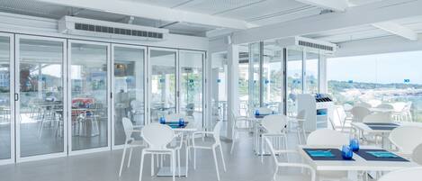 A modern indoor dining area with white furniture and glass partitions, offering views of the city and greenery.