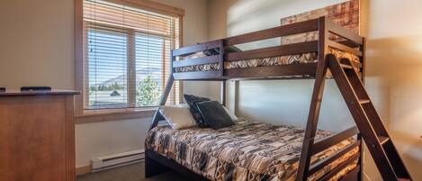 A bunk bed with a wooden frame and patterned bedding, a window with blinds, and a desk with a chair.