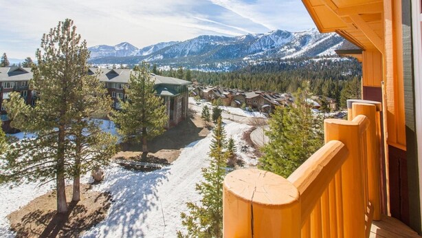 A wooden balcony with a view of a snow-covered landscape, a building with a green roof, and a forested area with mountains in the background. - Sunstone 329 (Mammoth Lakes)