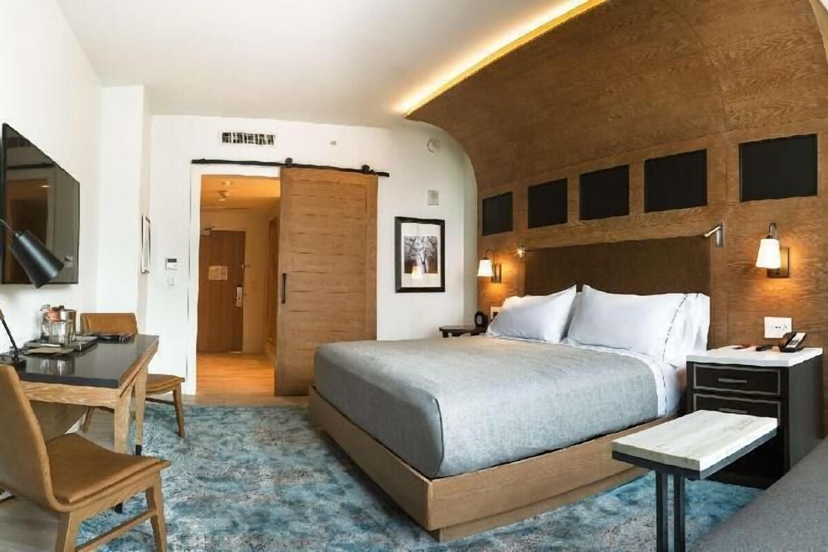 A hotel room with a bed, bedside table, lamp, sofa, desk with chair, and a TV.
