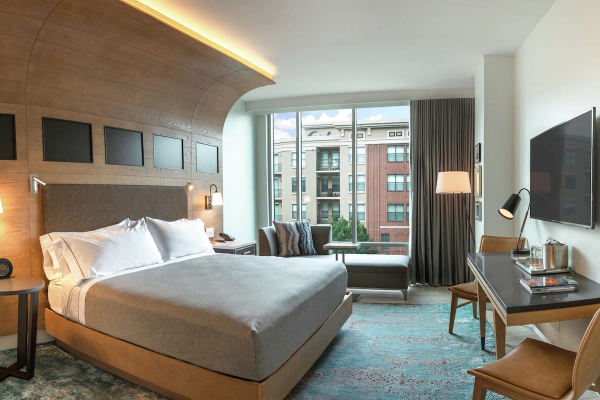 A hotel room with a large bed, a desk, a chair, a TV, and a view of buildings through the window.