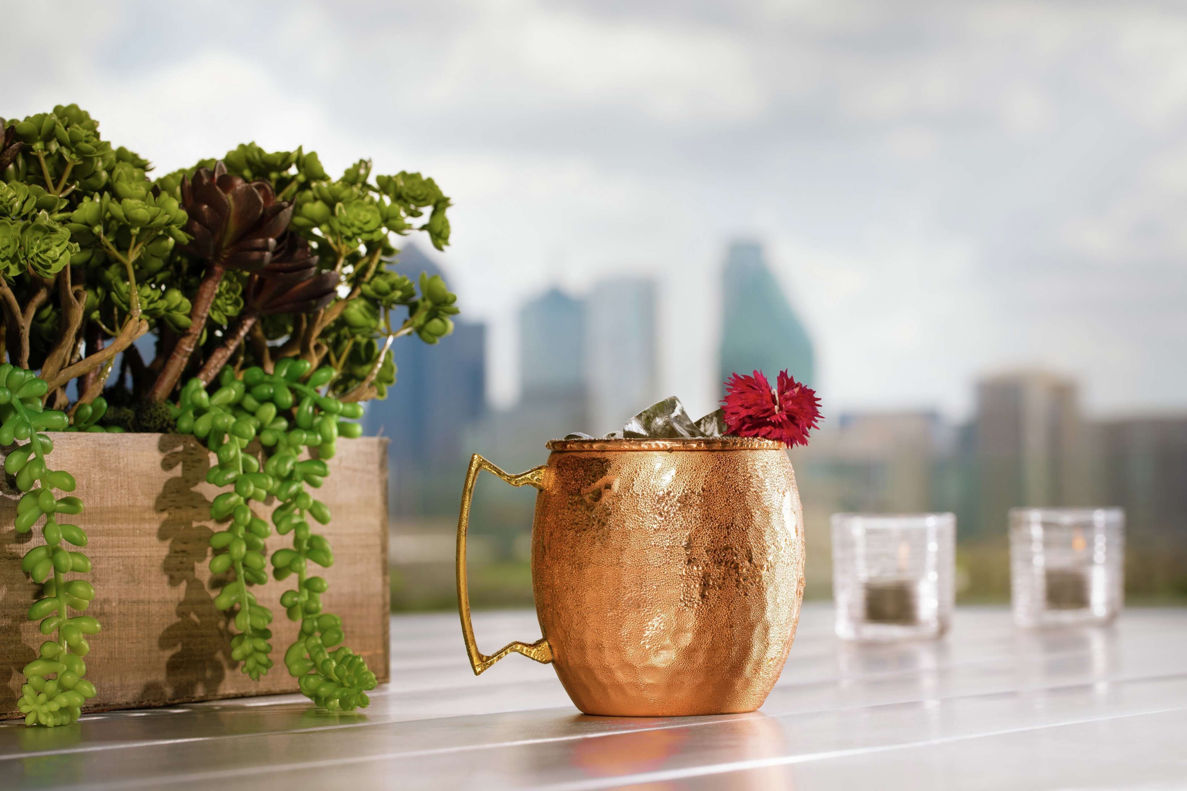 Una taza de cobre con asa sobre una mesa, una jardinera de madera con plantas verdes, y un paisaje urbano al fondo.