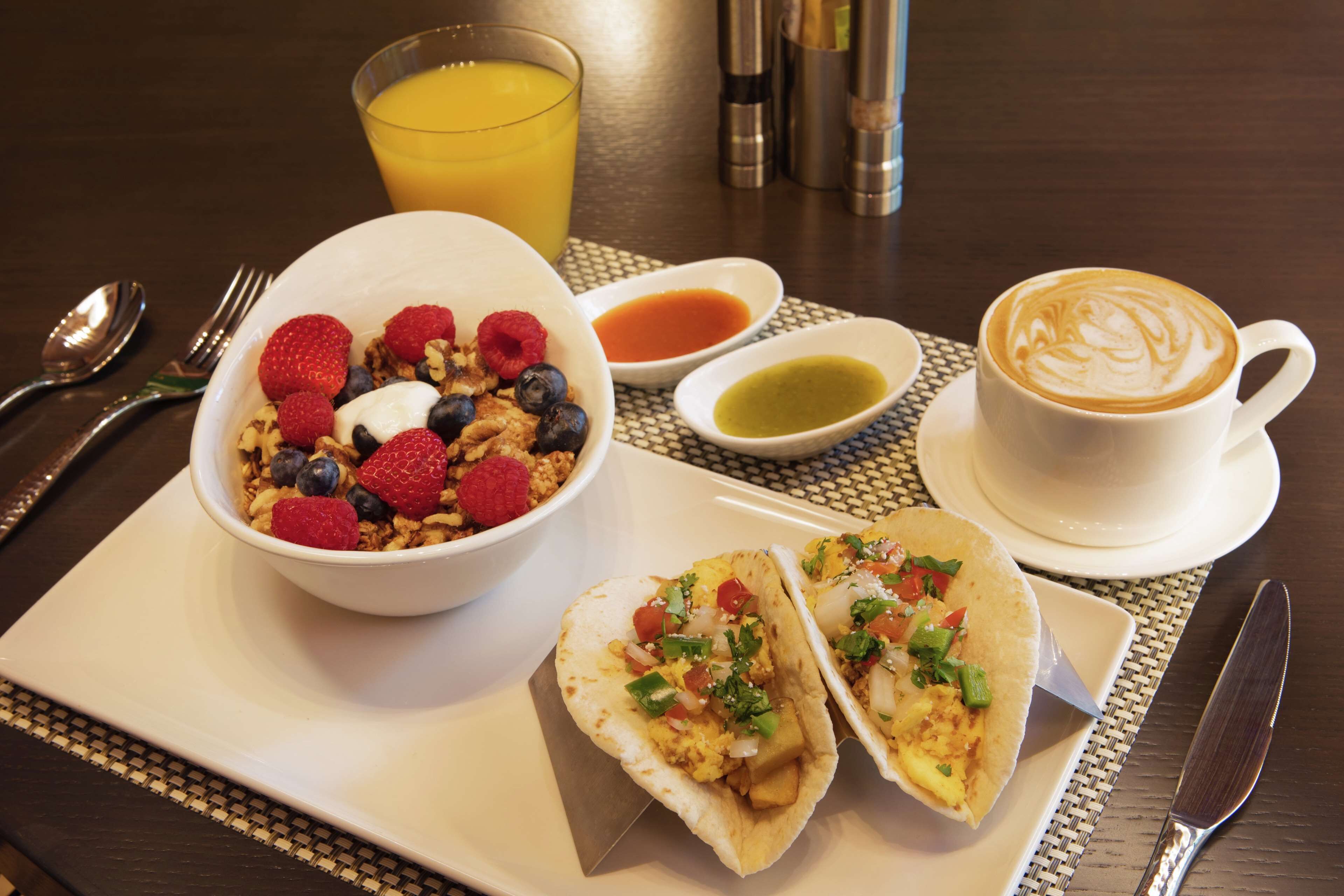 Un desayuno con un tazón de cereal cubierto de bayas, un vaso de jugo de naranja, dos tacos, una taza de café con arte latte y aderezos sobre una mesa.