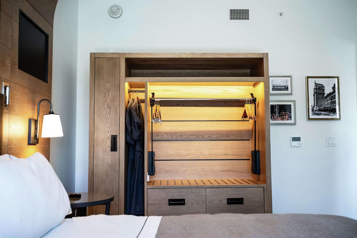 A hotel room with a wooden wardrobe, a bed, a nightstand, a lamp, and framed pictures on the wall.