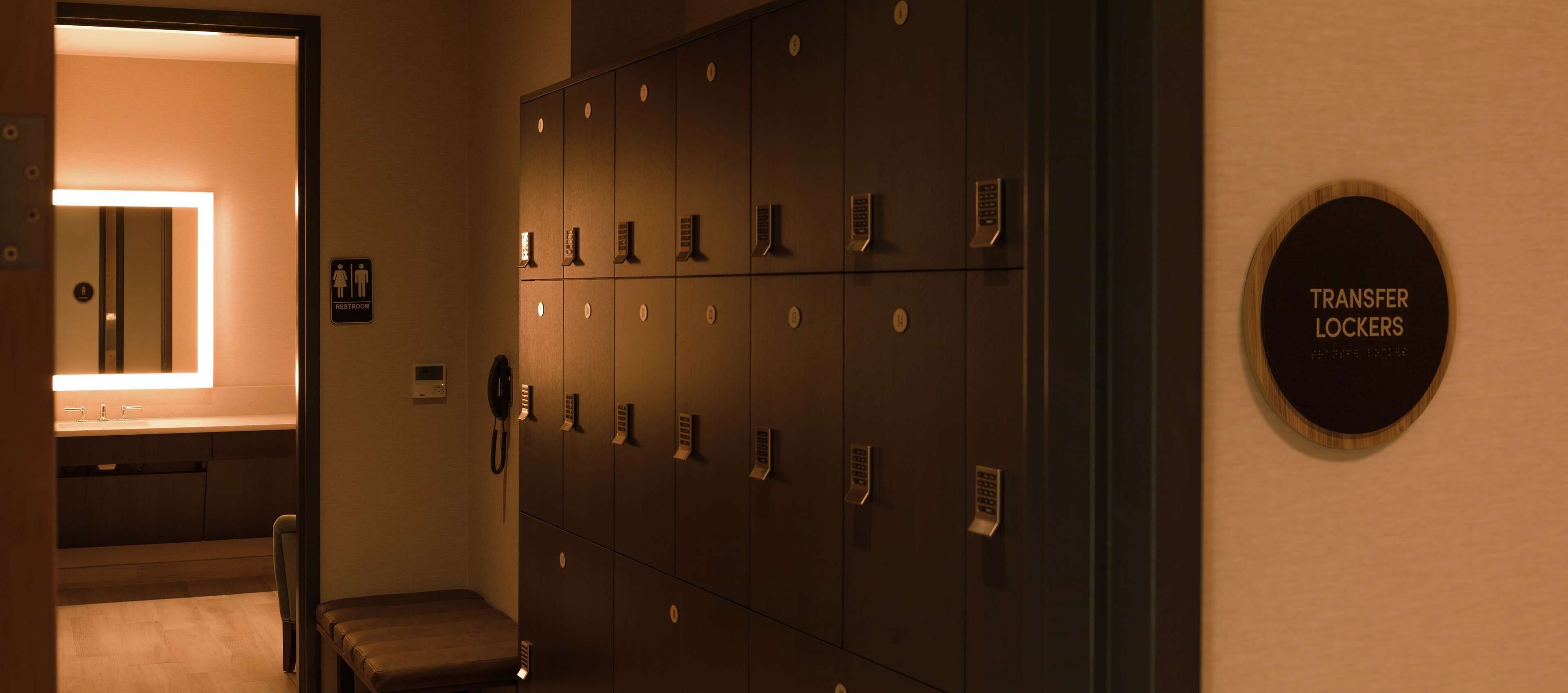 Een hotelkamer met een grote spiegel, een bankje en een rij kluisjes met de tekst 'TRANSFER KLUISJES'.