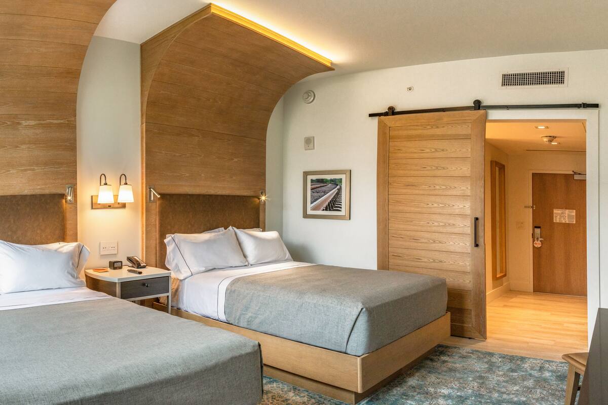 A hotel room with two beds, a wooden headboard, a nightstand, a lamp, and a door leading to another room.