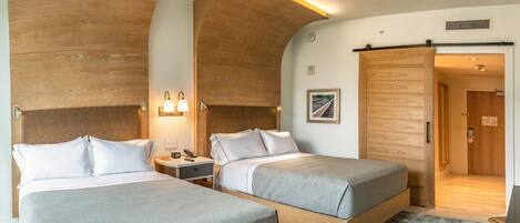 A hotel room with two beds, a wooden headboard, a nightstand, a lamp, and a door leading to another room.