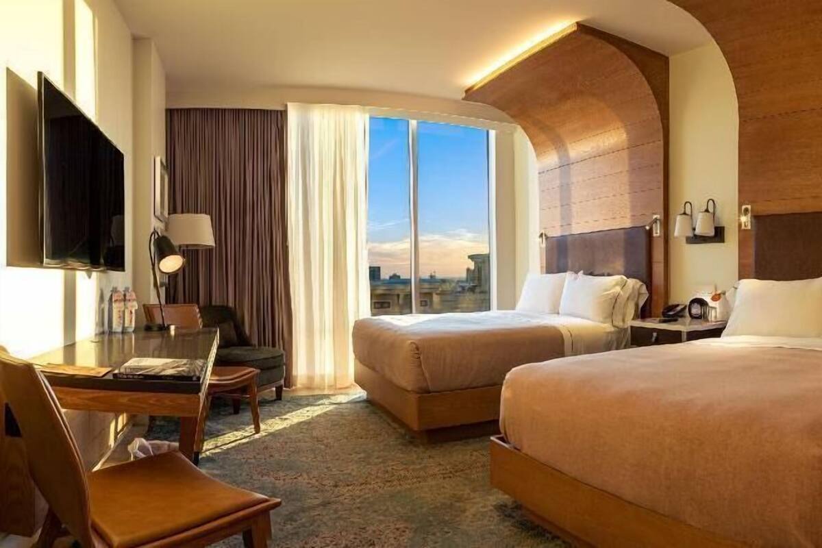 A hotel room with two beds, a desk, a chair, a TV, and a large window with a city view.