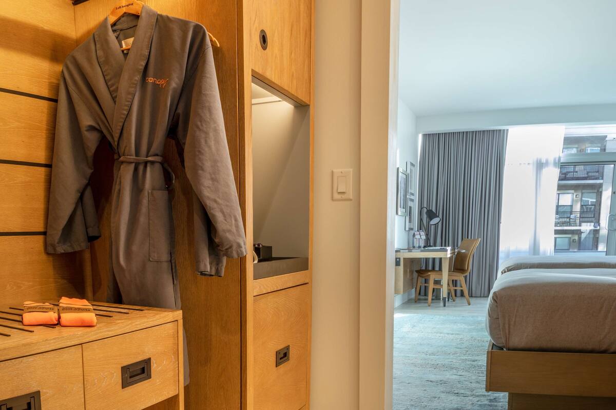 A hotel room with a wooden wardrobe, a robe hanging, a desk with a chair, and a bed.