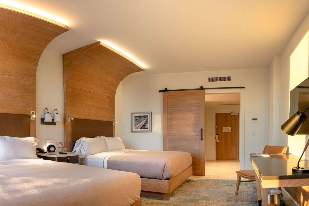 Habitación de hotel con dos camas, un escritorio, una silla, una lámpara, una mesita de noche y una lámpara de pared.