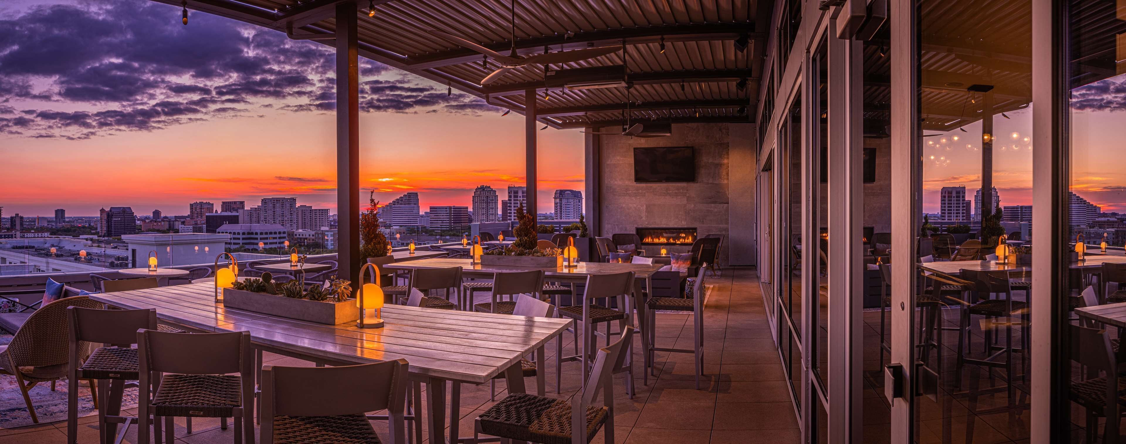 Un área de comedor al aire libre con mesas y sillas, un pozo de fuego y vistas a un horizonte urbano al atardecer.