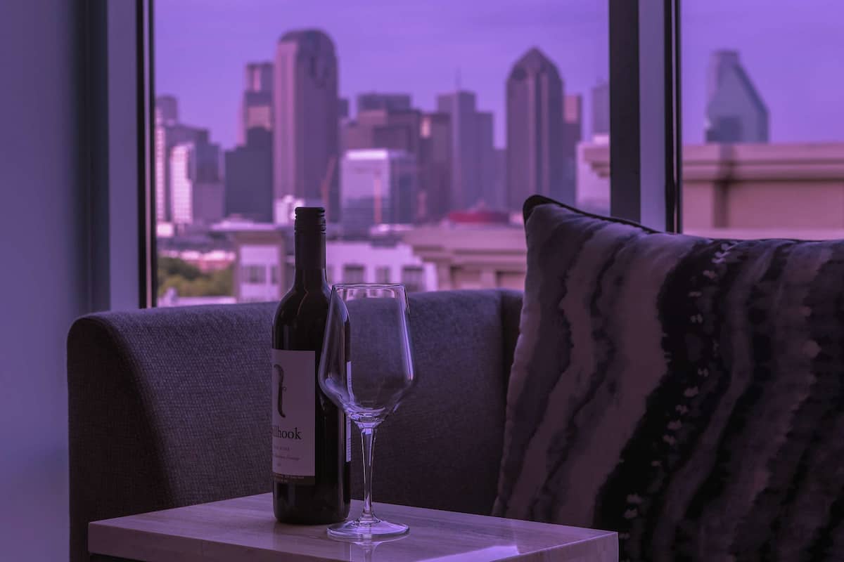 A bottle of wine and an empty glass on a table by a window with a cityscape view.