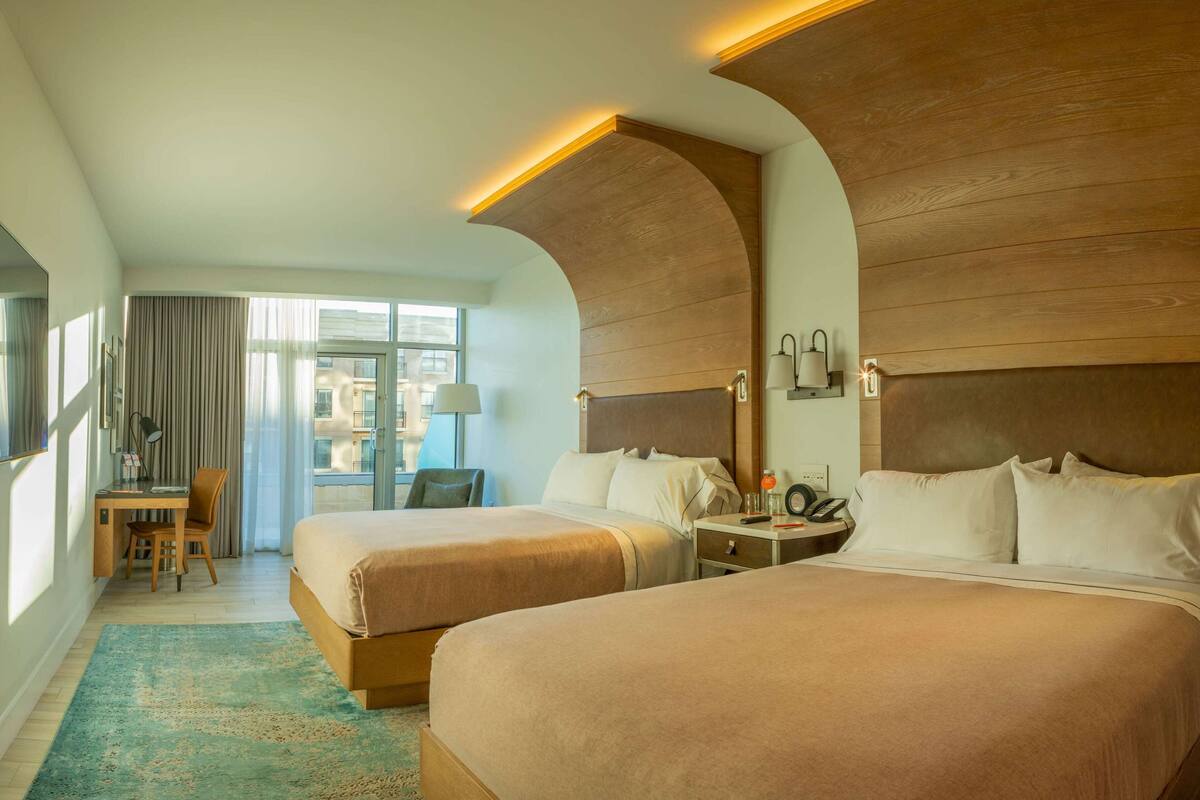 Quarto de hotel com duas camas, uma escrivaninha, uma cadeira, um abajur e vista para o exterior.
