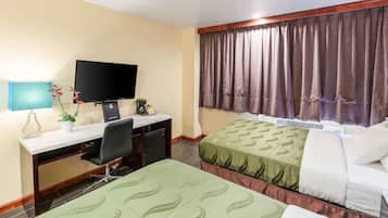 A hotel room with a desk, a television, two beds, a window with curtains, and a small plant on the desk.