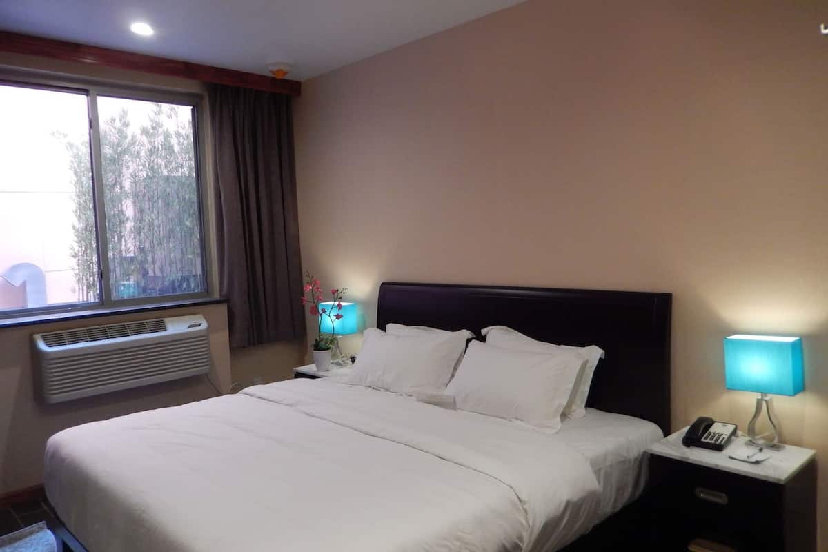 A hotel room with a bed, a bedside lamp, a nightstand with a phone, a window with curtains, and an air conditioning unit.