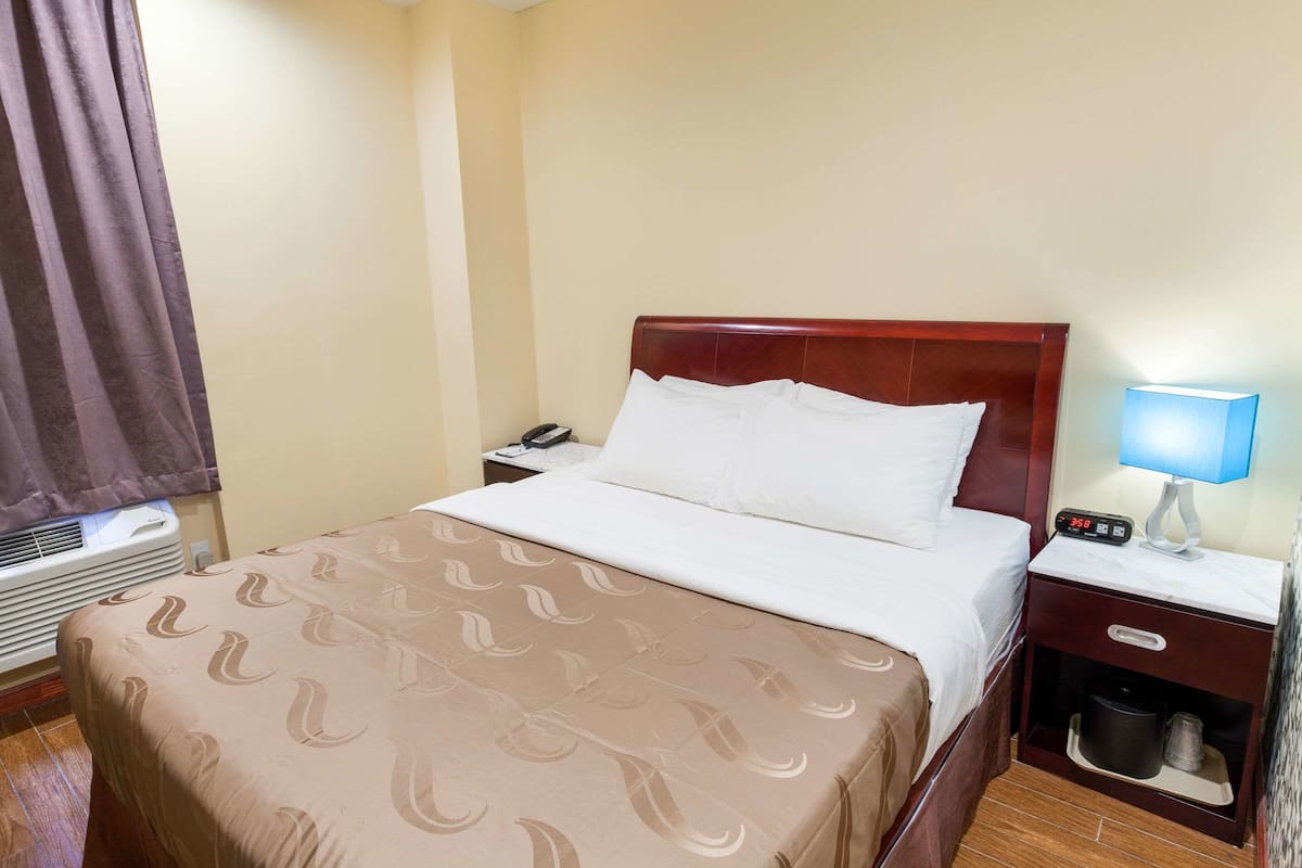 A hotel room with a bed, a nightstand with a lamp, a phone, and a trash bin.