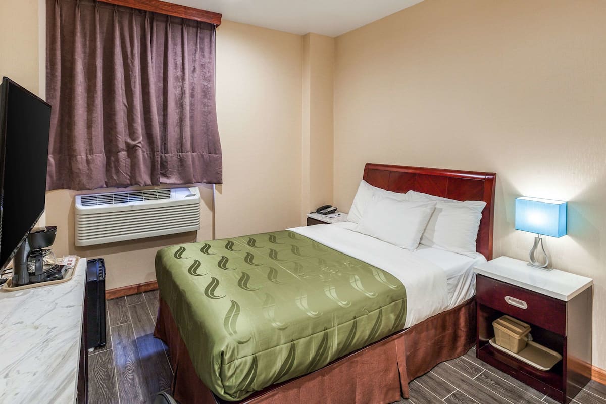 A hotel room with a bed, a nightstand, a lamp, an air conditioning unit, a TV, and a window with curtains.