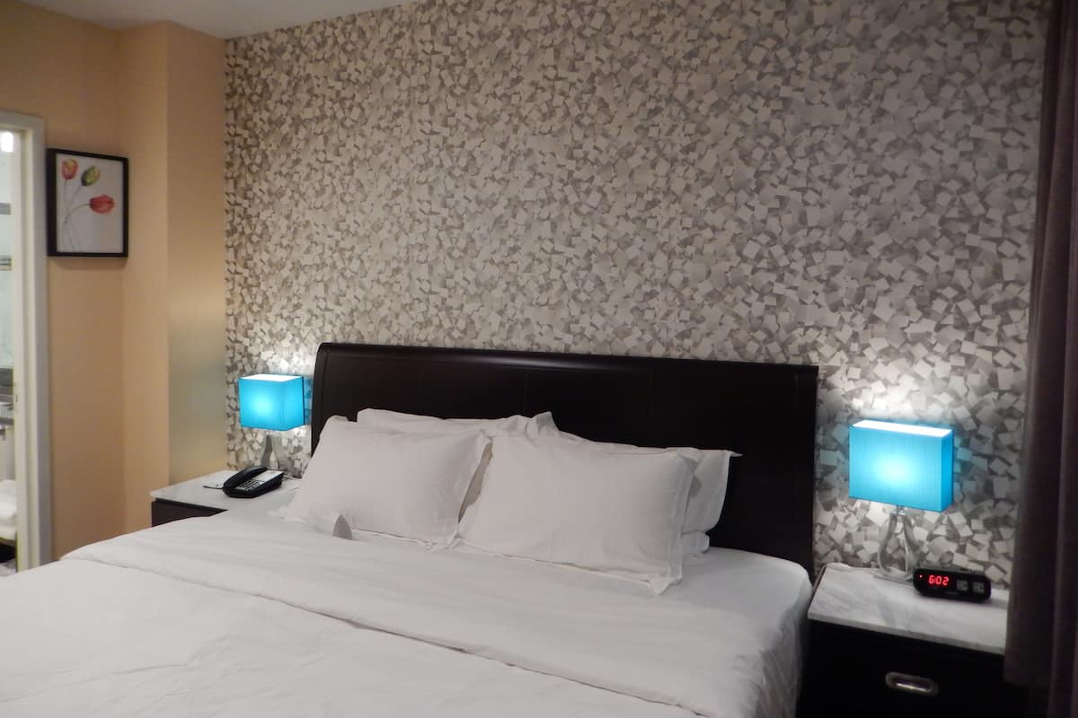 A hotel room with a bed, two bedside lamps, a nightstand with a phone, and a digital clock displaying 6:02.