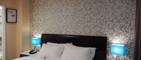 Habitación de hotel con una cama, dos lámparas de noche, una mesita de noche con un teléfono y un reloj digital que marca las 6:02.