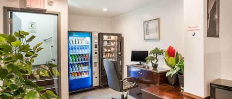A reception area with a desk, a computer, a microwave sign, a vending machine, and a staircase leading to an elevator.