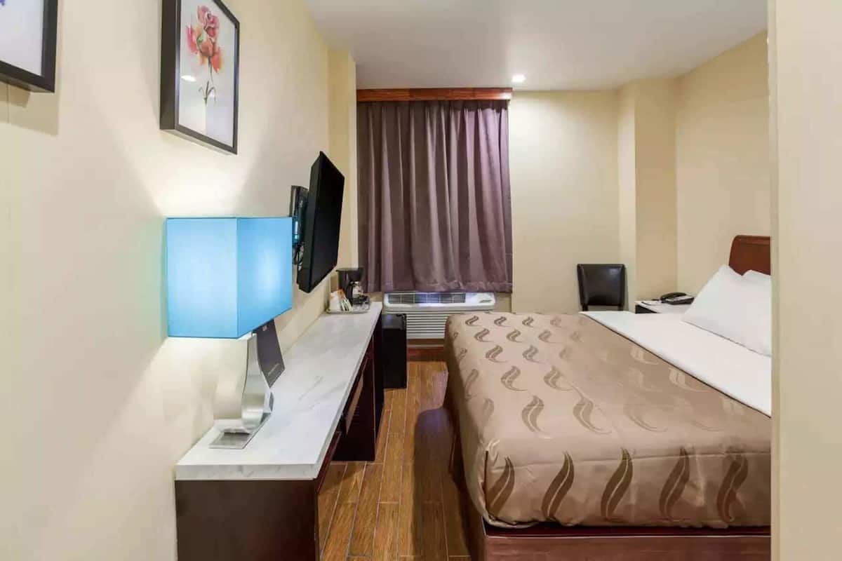 A hotel room with a bed, a desk with a computer, a TV, and a window with curtains.