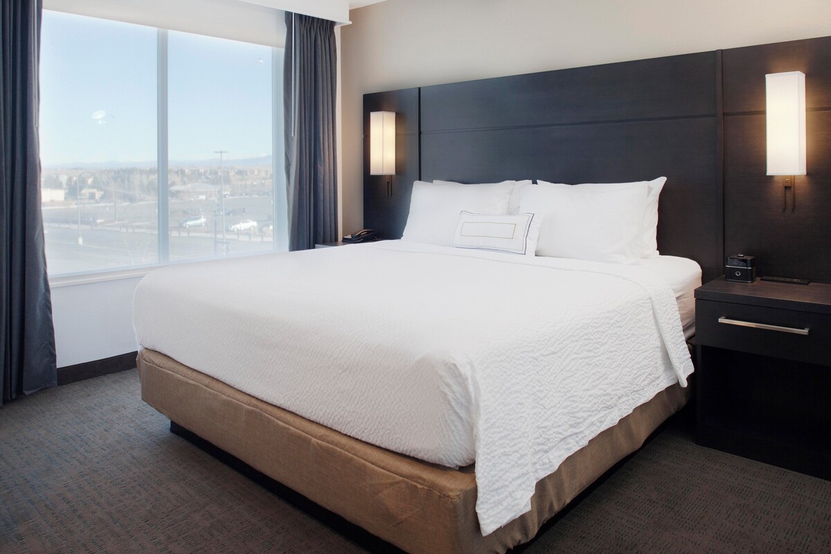 A large bed with a headboard, two bedside tables, and a view of a cityscape through a large window.