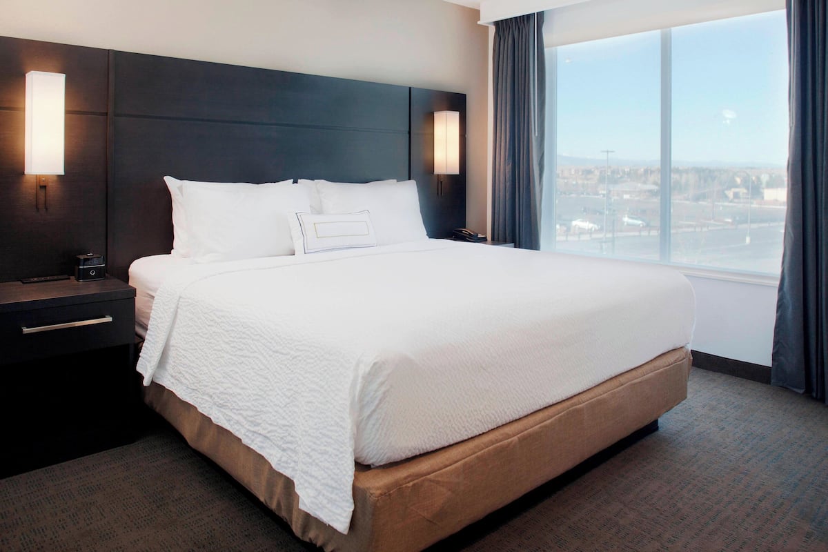 A hotel room with a large bed, dark headboard, bedside lamps, and a view of the city through the window.