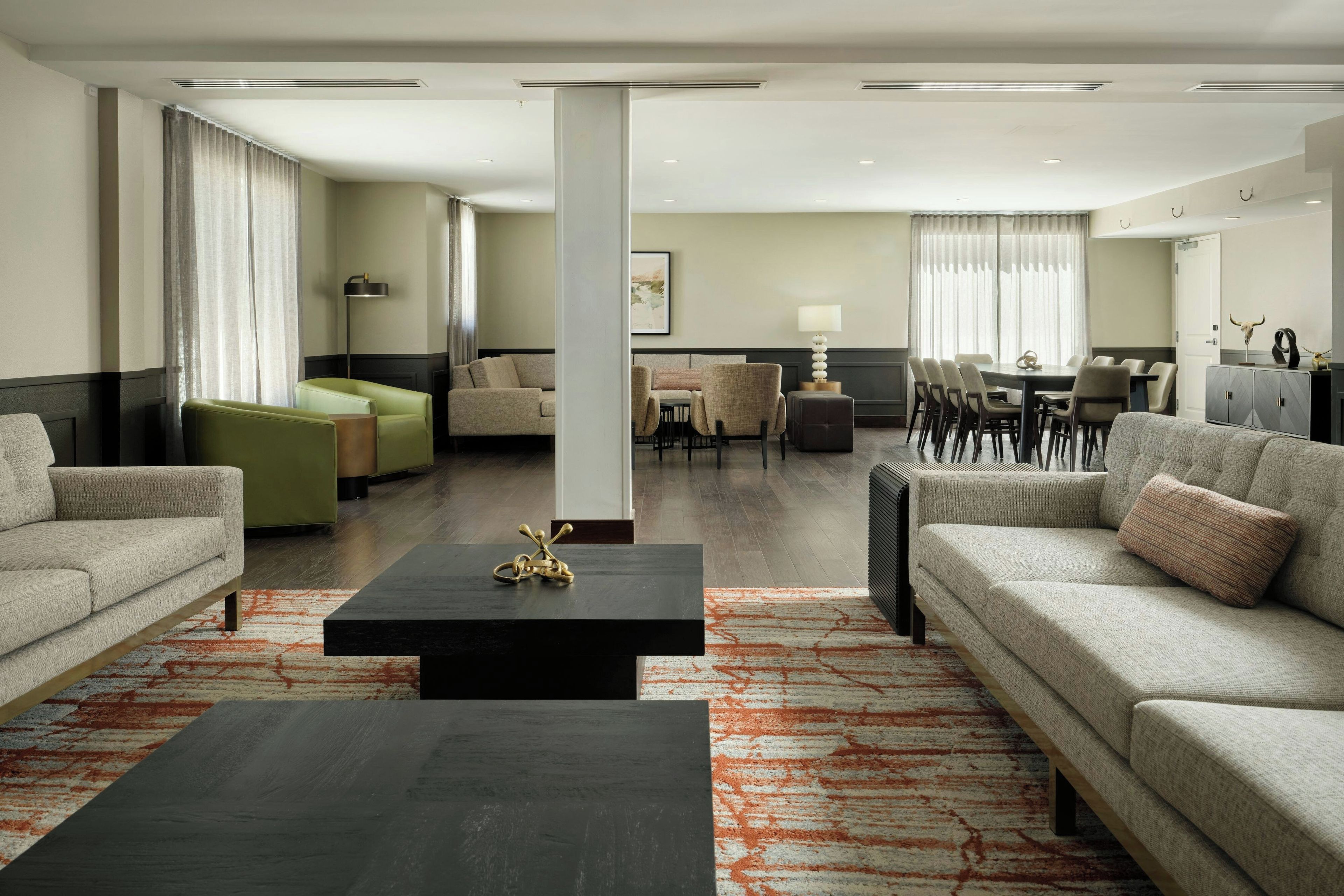 Une chambre d’hôtel moderne avec un canapé, un fauteuil vert, une table à manger avec des chaises et une grande fenêtre avec des rideaux.