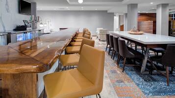 A modern hotel conference room with a long wooden table, several chairs, a bar area, and a television mounted on the wall.