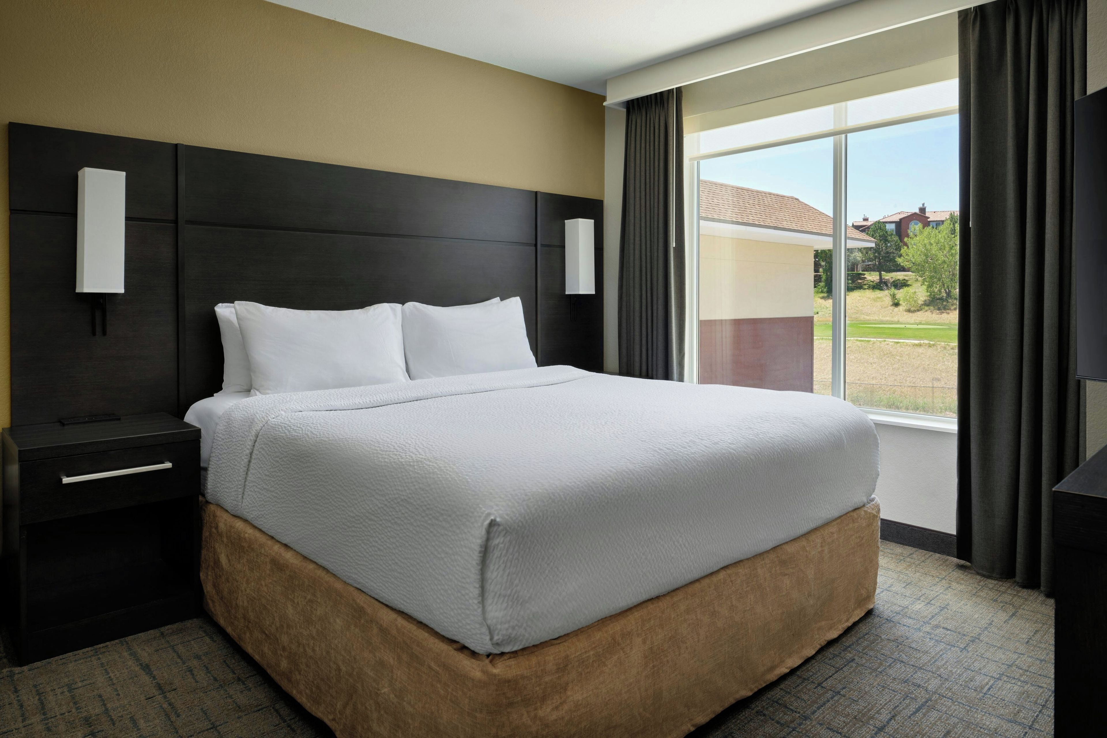 A hotel room with a large bed, a nightstand, a bedside table, a window with curtains, and a view of the outdoors.