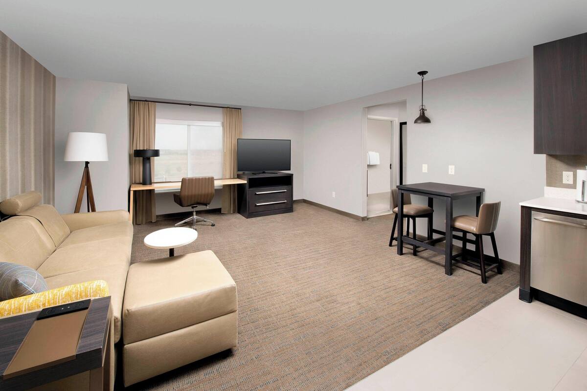A modern hotel room with a sofa, a dining table with chairs, a TV, and a kitchenette.