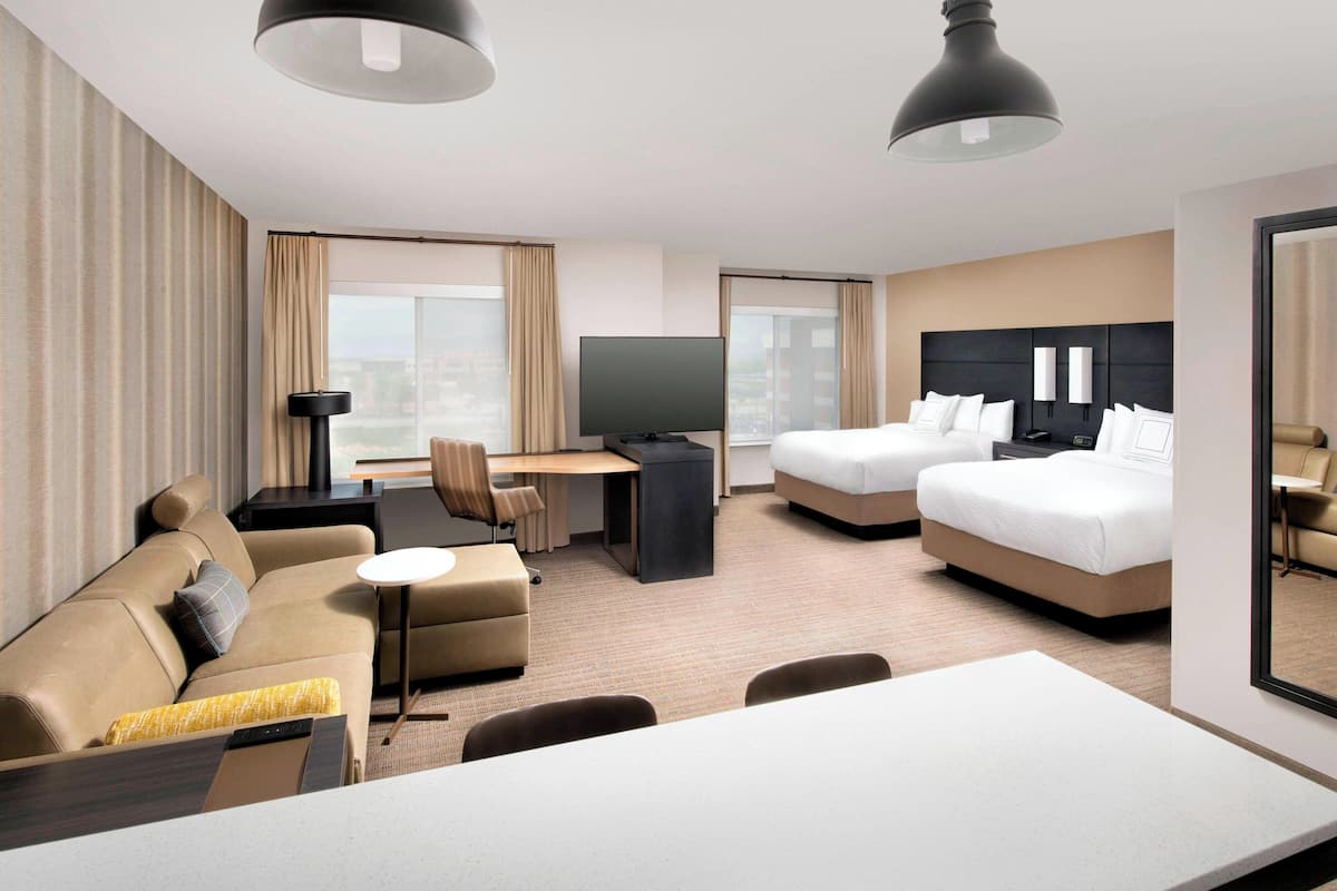 A modern hotel room with a sofa, two single beds, a desk with a TV, and a large window offering a city view.