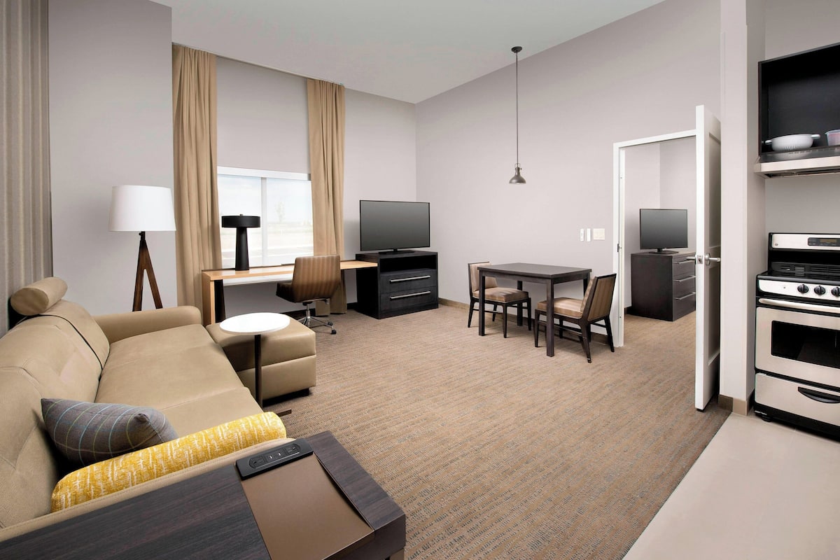 A modern hotel room with a sofa, a dining table, chairs, a TV, a lamp, and a kitchen area with an oven.