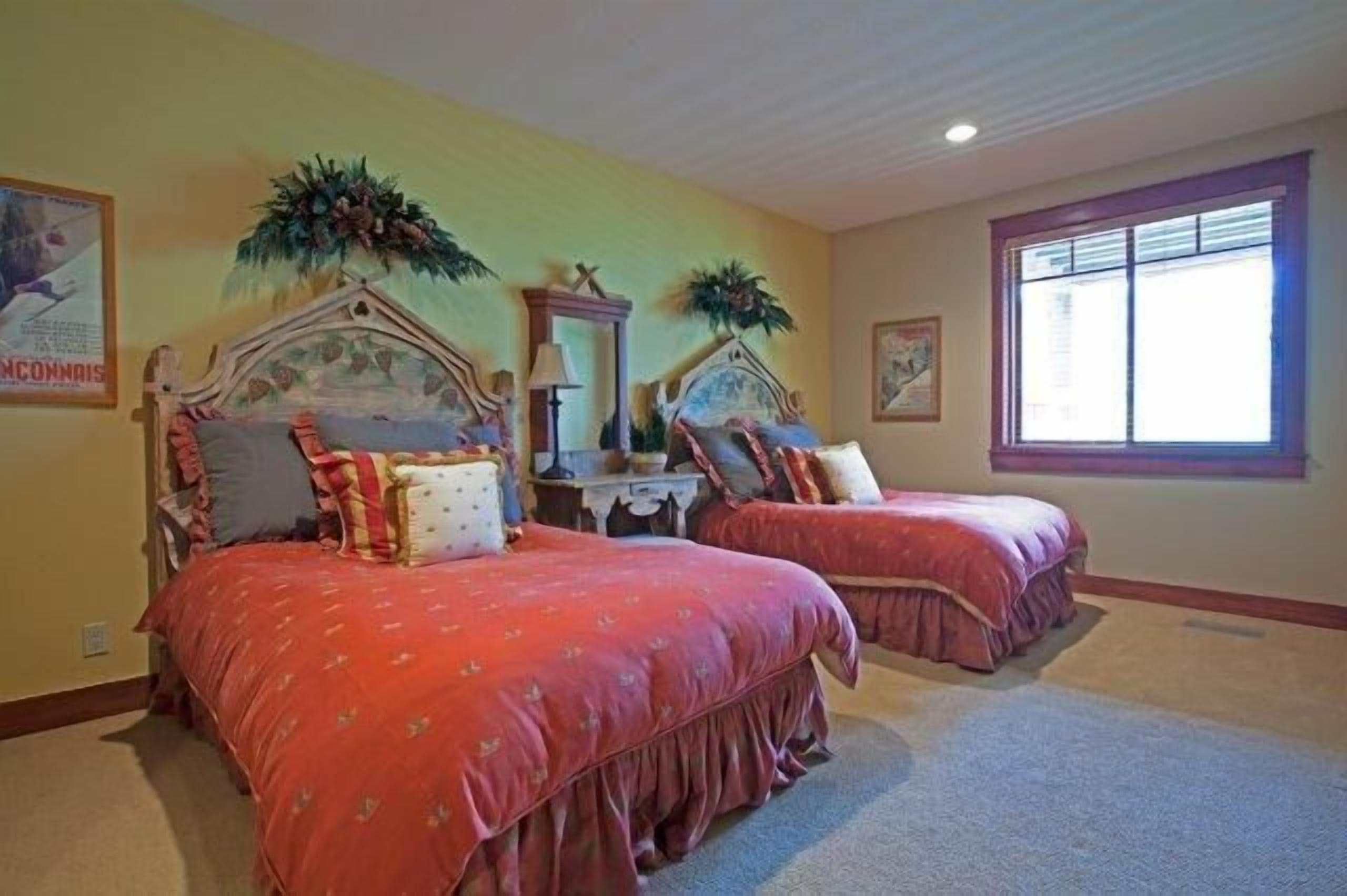 A bedroom with two beds, each with a headboard and pillows. There is a window with blinds, a wall-mounted mirror, and a framed picture.