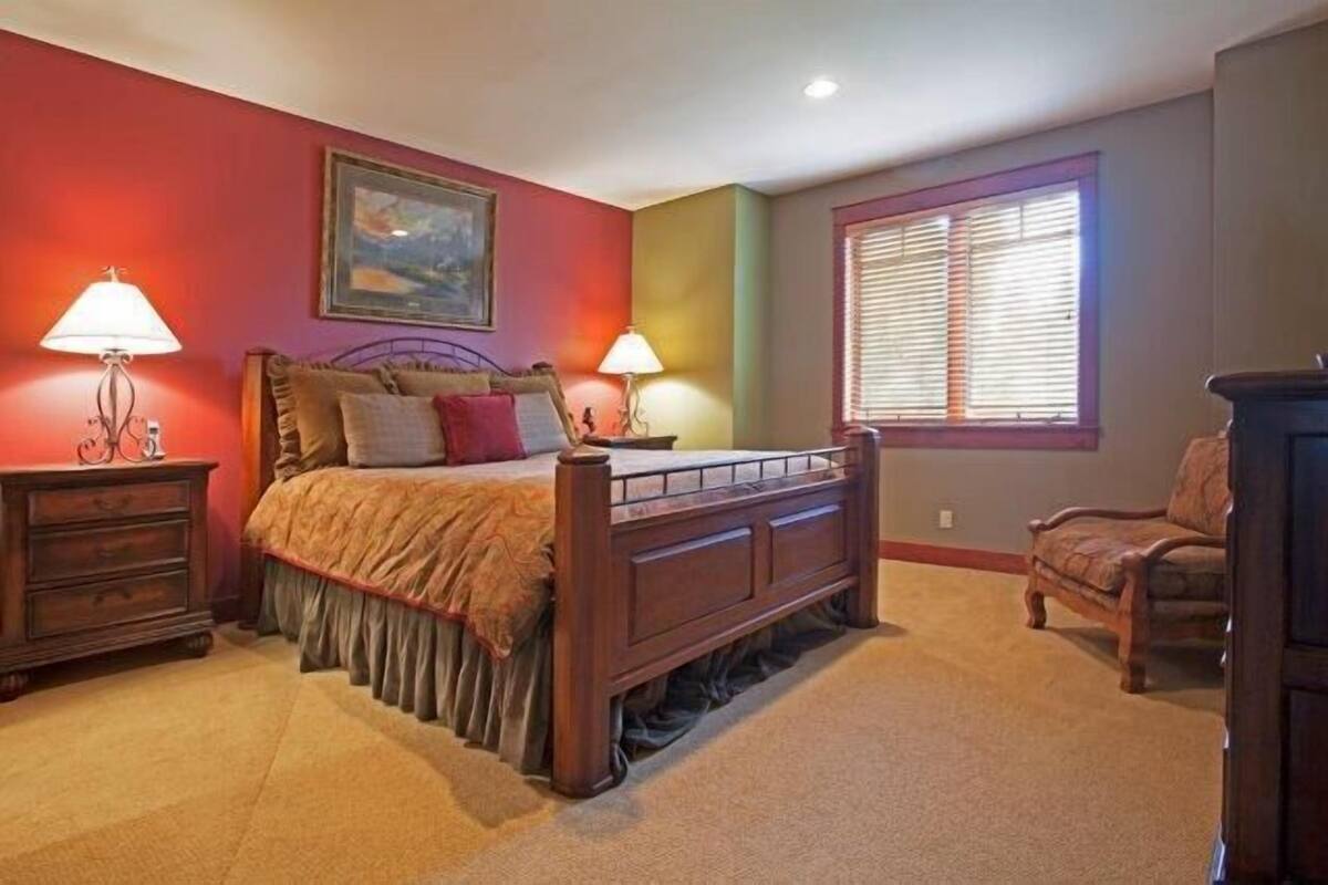 A bedroom with a wooden bed, two bedside tables with lamps, a chair, and a window with blinds.