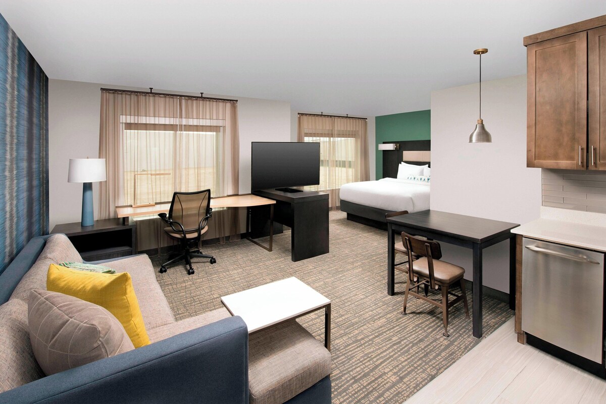 Habitación de hotel con sofá, escritorio y silla, cama, televisión, mesita y kitchenette.