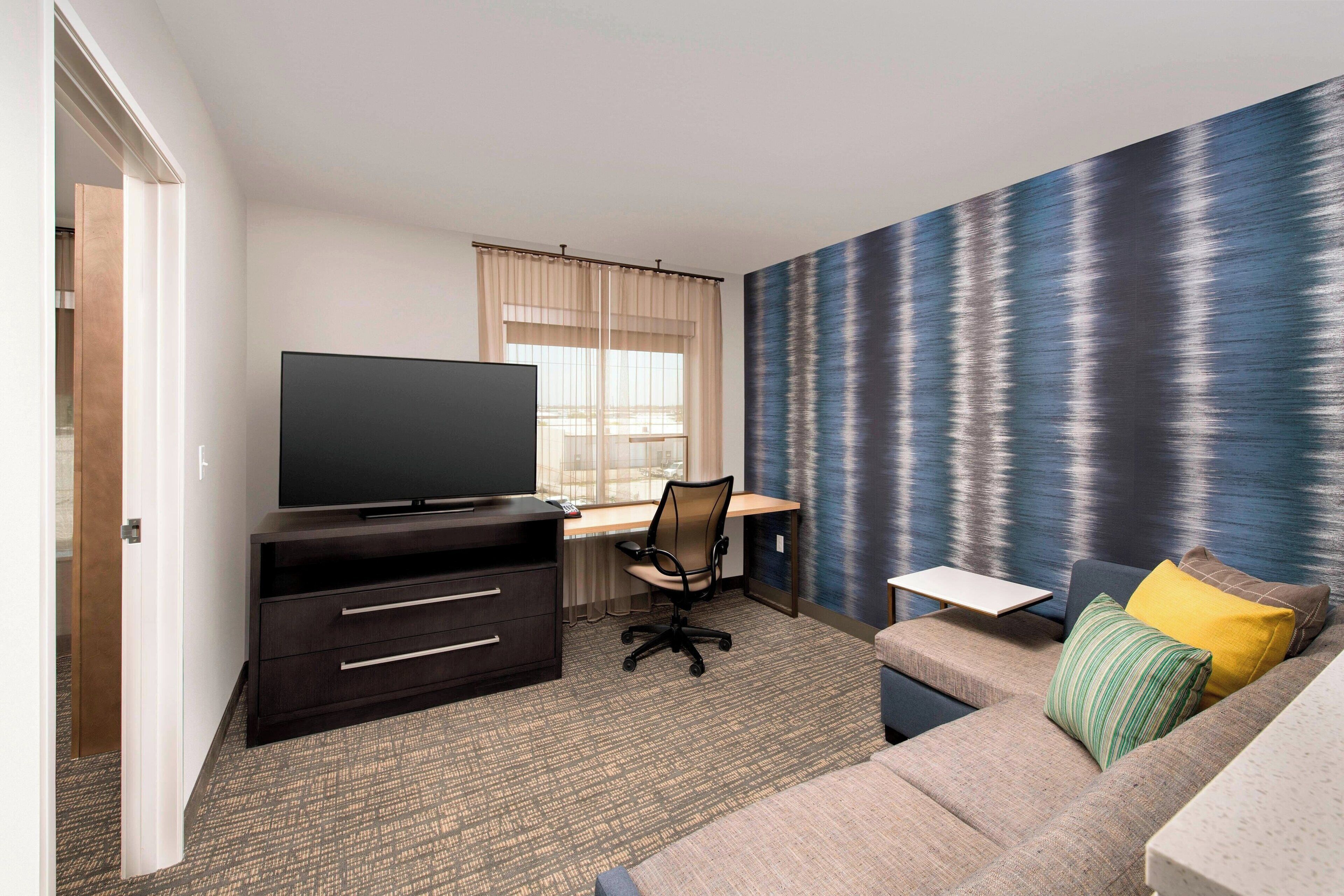 Une chambre d’hôtel équipée d’une télévision à écran plat, d’un bureau avec une chaise, d’un canapé avec des coussins et d’une grande fenêtre avec des rideaux.