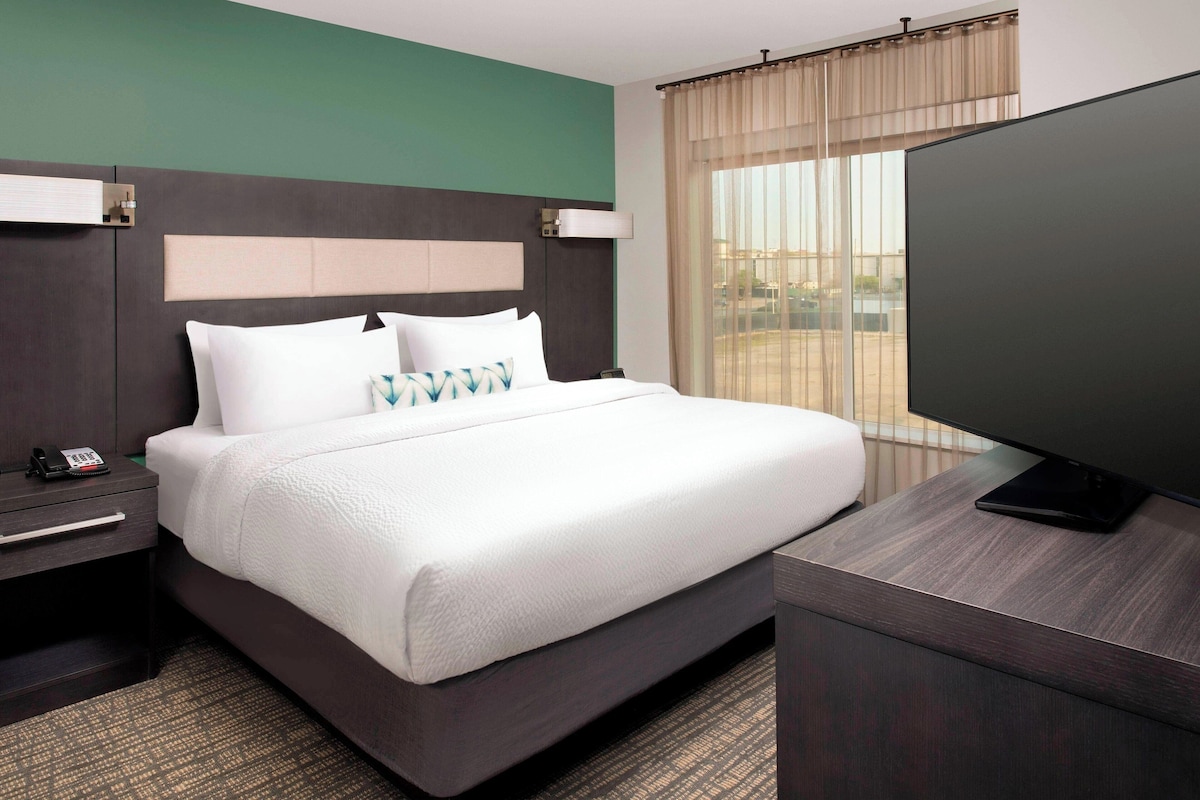 Habitación de hotel con una cama grande, una mesita de noche con teléfono, un televisor de pantalla plana sobre un soporte y una ventana con cortinas.