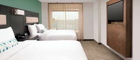 A hotel room with two beds, a large flat-screen TV mounted on the wall, a window with sheer curtains, and a carpeted floor.