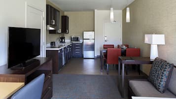 A compact hotel room with a kitchenette, a flat-screen TV, a sofa, a dining table, and a patterned rug.